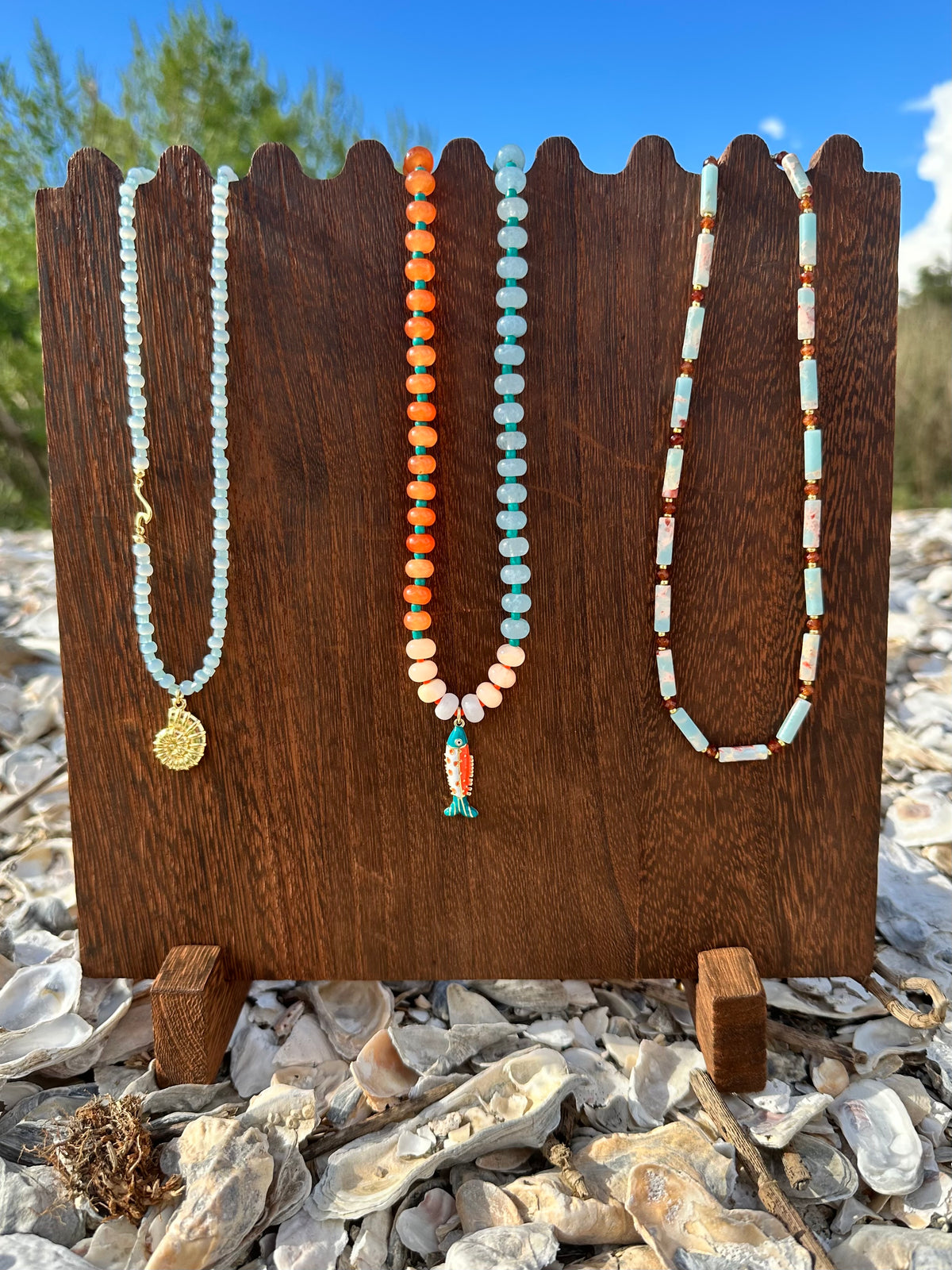Three beaded necklaces displayed on a dark wooden stand outdoors on a rocky surface with shells and greenery in the background. The necklaces feature light blue beads with a gold medallion, orange and cream beads with a small fish charm, and cream and light blue beads.