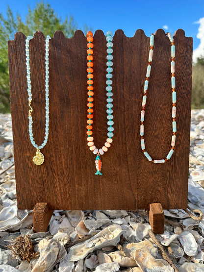 Three beaded necklaces displayed on a dark wooden stand outdoors on a rocky surface with shells and greenery in the background. The necklaces feature light blue beads with a gold medallion, orange and cream beads with a small fish charm, and cream and light blue beads.