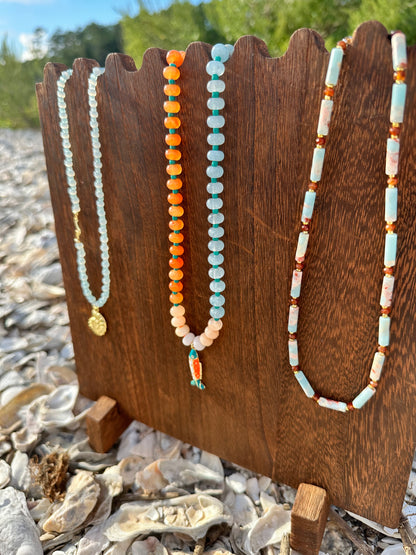 Three beaded necklaces displayed on a rustic wooden stand outdoors on a rocky beach. The necklaces feature light blue, orange, peach, and white beads with small gold and coral accents.