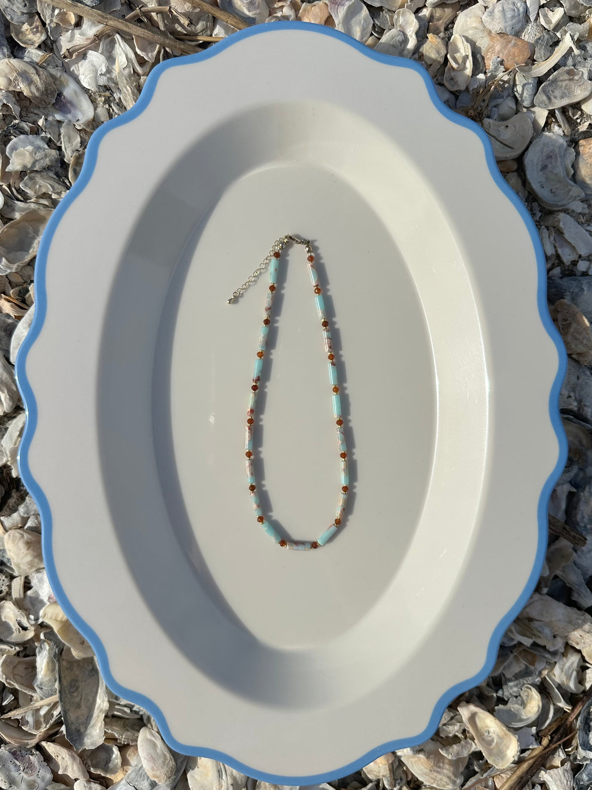 A beaded necklace with light blue and orange beads with gold accents, displayed on a white oval platter with a blue rim, set against a background of small seashells.