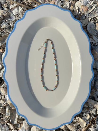 A beaded necklace with light blue and orange beads with gold accents, displayed on a white oval platter with a blue rim, set against a background of small seashells.