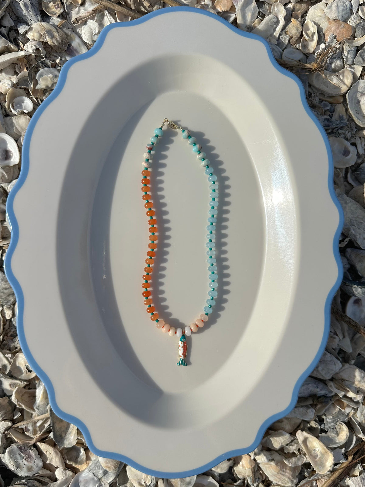 A beaded necklace with orange, light blue, and peach beads, displayed on a white oval platter with a blue rim set against a background of small seashells.