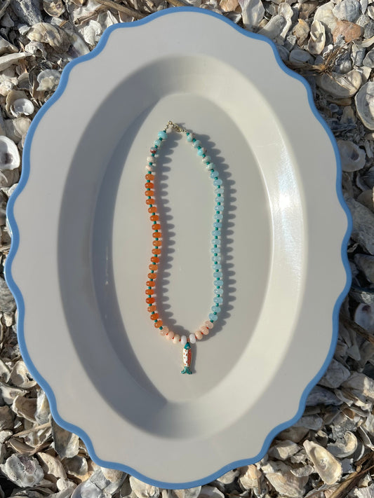 A beaded necklace with orange, light blue, and peach beads, displayed on a white oval platter with a blue rim set against a background of small seashells.