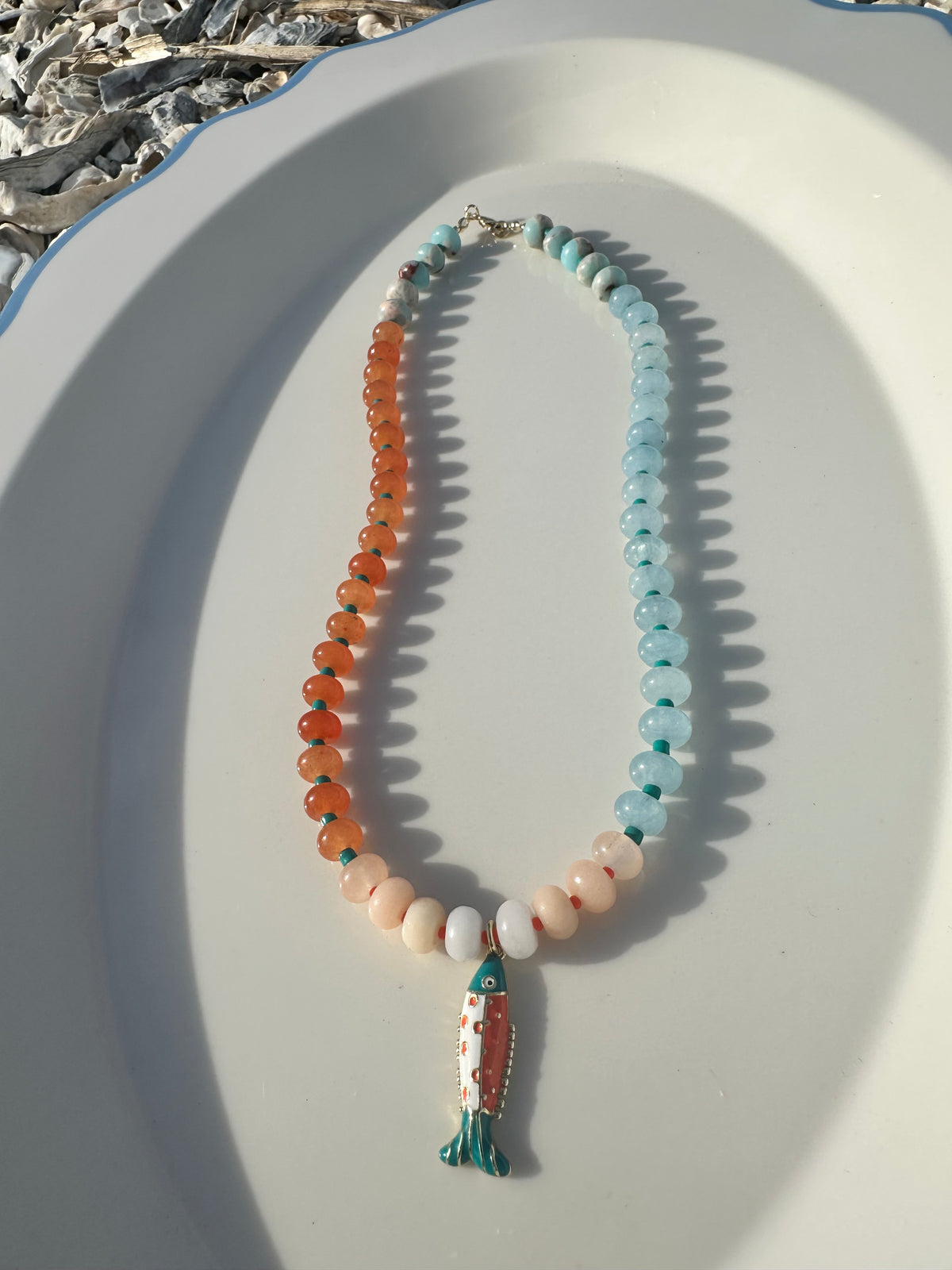A beaded necklace with orange and light blue beads, teal spacer beads, and a fish pendant, displayed on a white oval tray.
