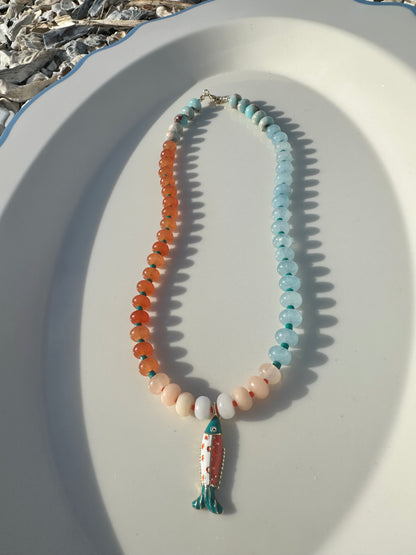 A beaded necklace with orange and light blue beads, teal spacer beads, and a fish pendant, displayed on a white oval tray.