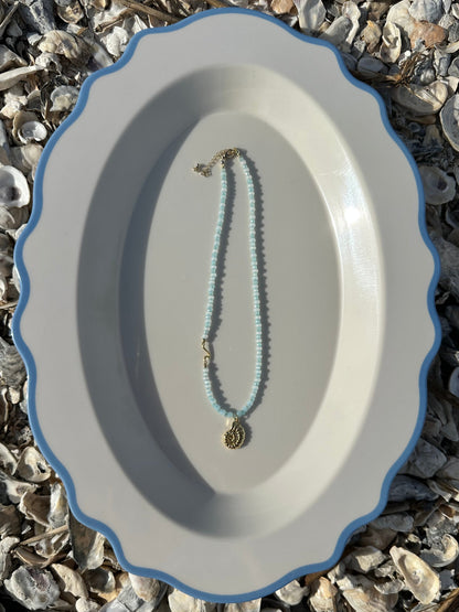 A delicate beaded necklace with light blue beads and a gold-toned pendant, displayed on a white oval plate with a blue rim, set against a background of small seashells.