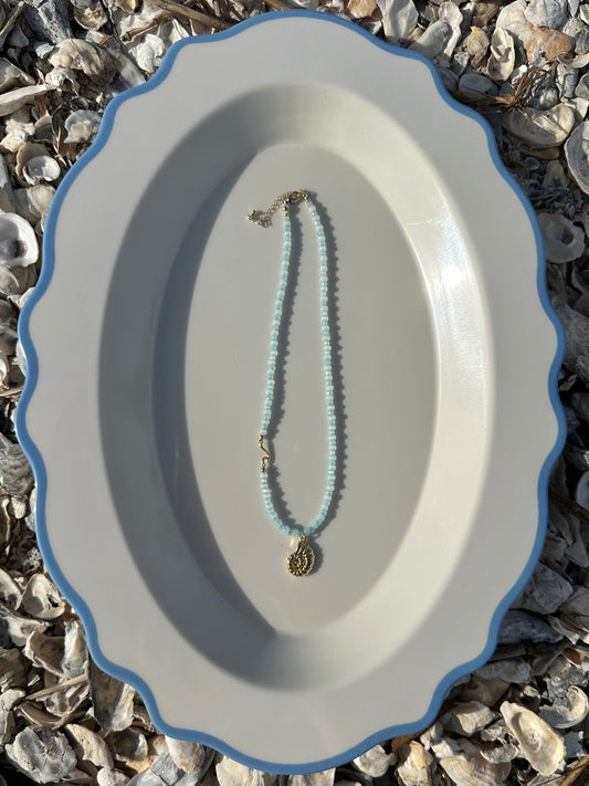 A delicate beaded necklace with light blue beads and a gold-toned pendant, displayed on a white oval plate with a blue rim, set against a background of small seashells.