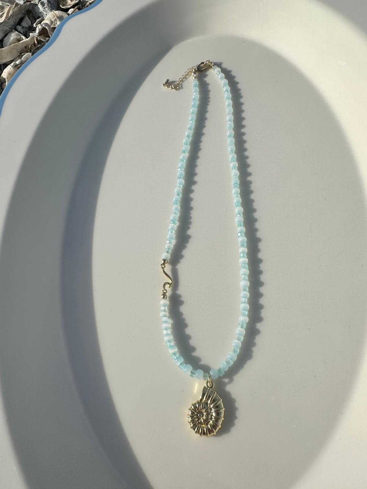 A delicate necklace with light blue beads and a gold-toned seashell pendant, displayed on a white oval tray.