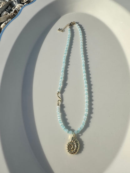 A delicate necklace with light blue beads and a gold-toned seashell pendant, displayed on a white oval tray.