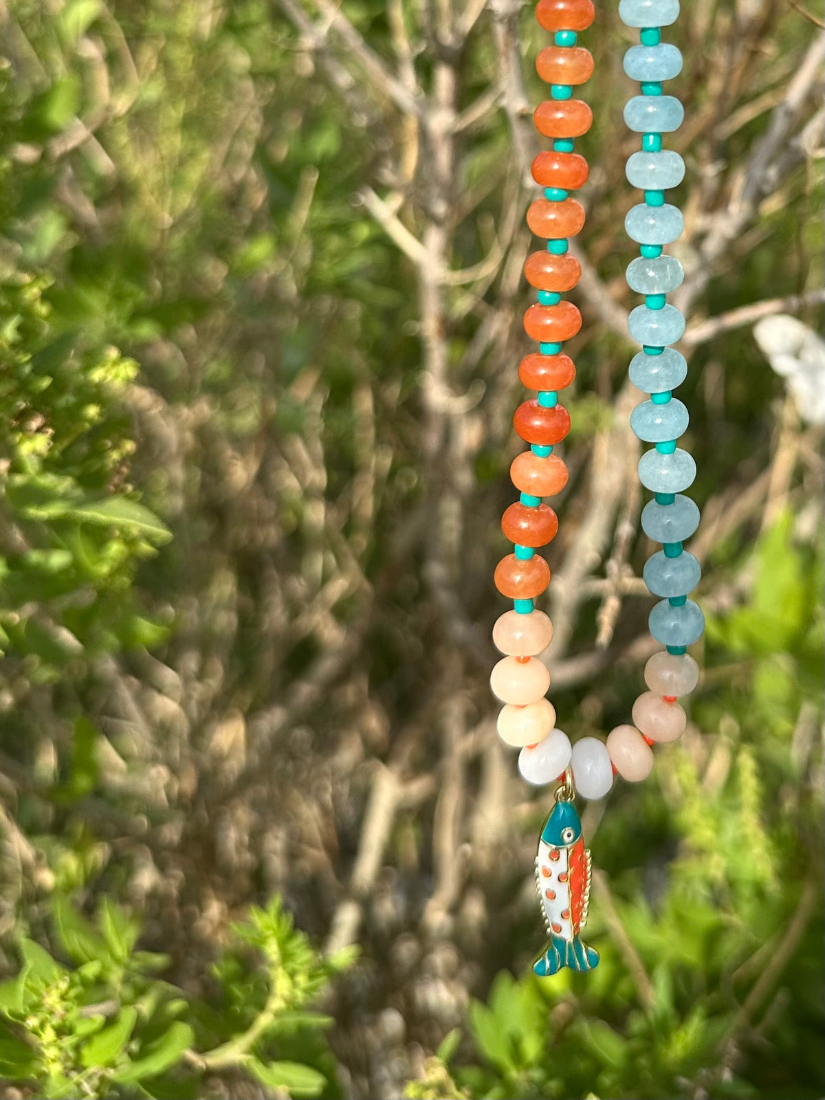 A beaded necklace with two strands hanging outdoors against a blurred background of green foliage and branches. One strand features round orange beads, the other light blue beads, and both transition through peach, cream, and white beads.