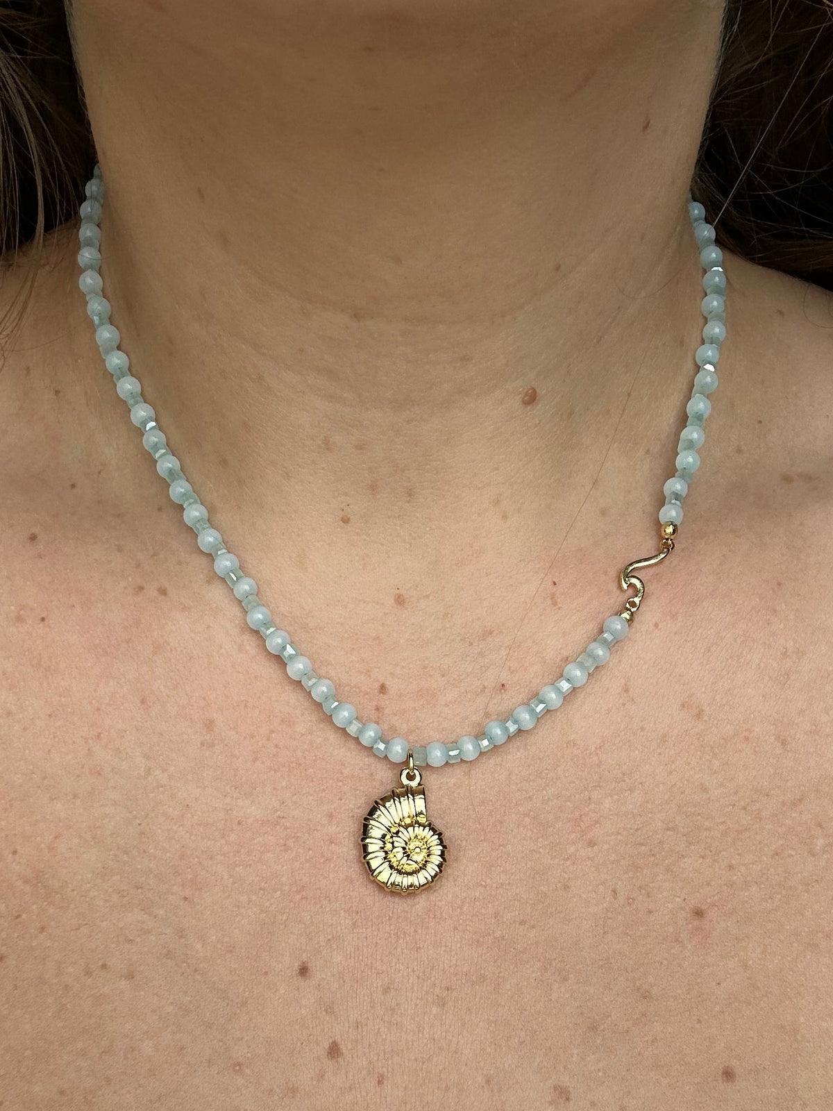 A close-up of a woman's neck and chest wearing a light blue beaded necklace with a gold spiral shell pendant.