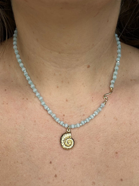 A close-up of a woman's neck and chest wearing a light blue beaded necklace with a gold spiral shell pendant.