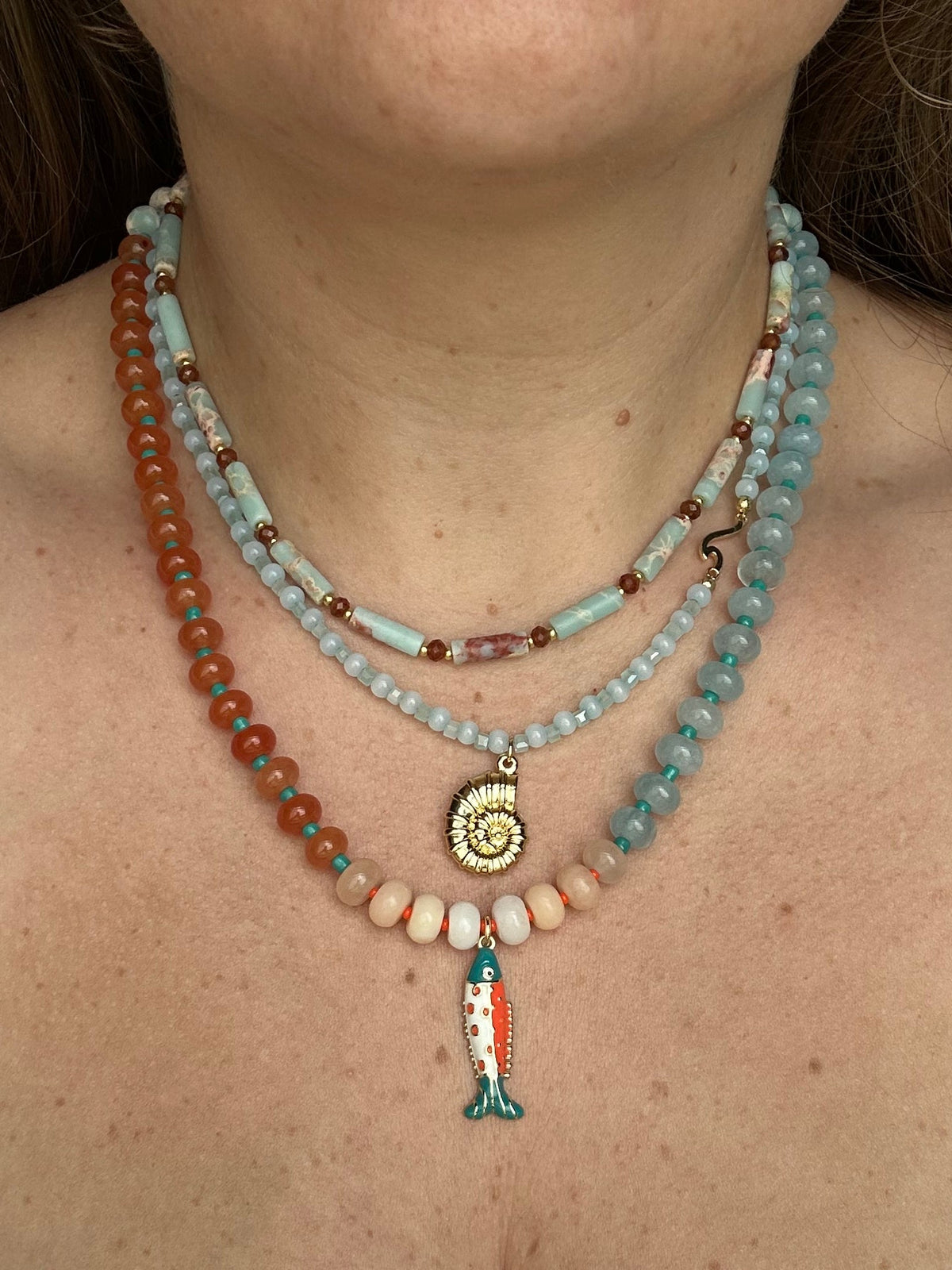 A close-up of a woman wearing a multi-strand bohemian beaded necklace set. The necklace features light blue turquoise beads, orange-red beads, cream beads, a gold spiral shell pendant, and a teal and orange fish charm.