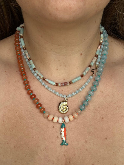 A close-up of a woman wearing a multi-strand bohemian beaded necklace set. The necklace features light blue turquoise beads, orange-red beads, cream beads, a gold spiral shell pendant, and a teal and orange fish charm.
