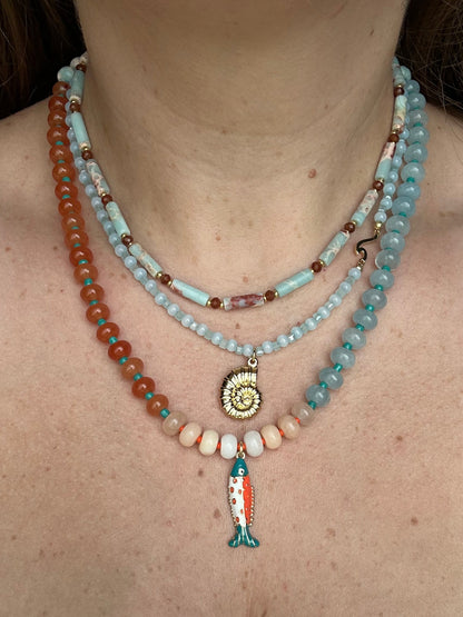 A close-up of a person's neck and upper chest wearing three layered beaded necklaces. The necklaces include light blue, orange, and cream beads with a gold spiral shell pendant and a small fish charm.