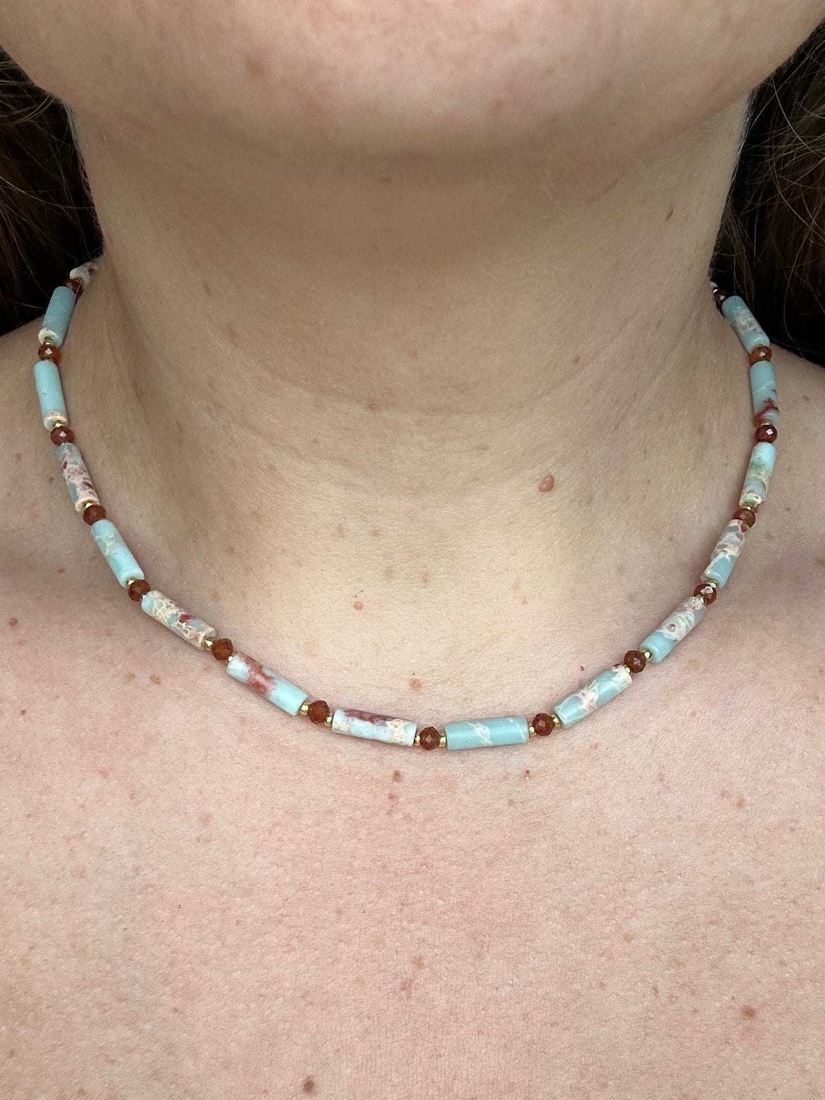 A close-up of a person's neck and upper chest wearing a beaded necklace made of light blue, white, and dark orange beads.