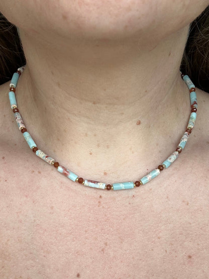 A close-up of a person's neck and upper chest wearing a beaded necklace made of light blue, white, and dark orange beads.