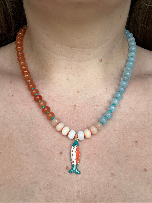 A close-up of a person's neck and chest wearing a beaded necklace. The necklace features a mix of orange, light blue, and cream beads, with a small fish-shaped pendant in the center.